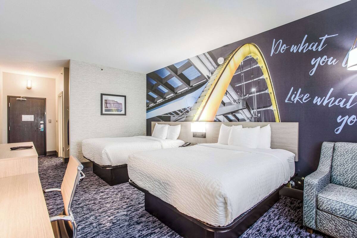 A hotel room with two beds, a desk, and a chair. There is a large wall mural with a quote and a yellow arch.