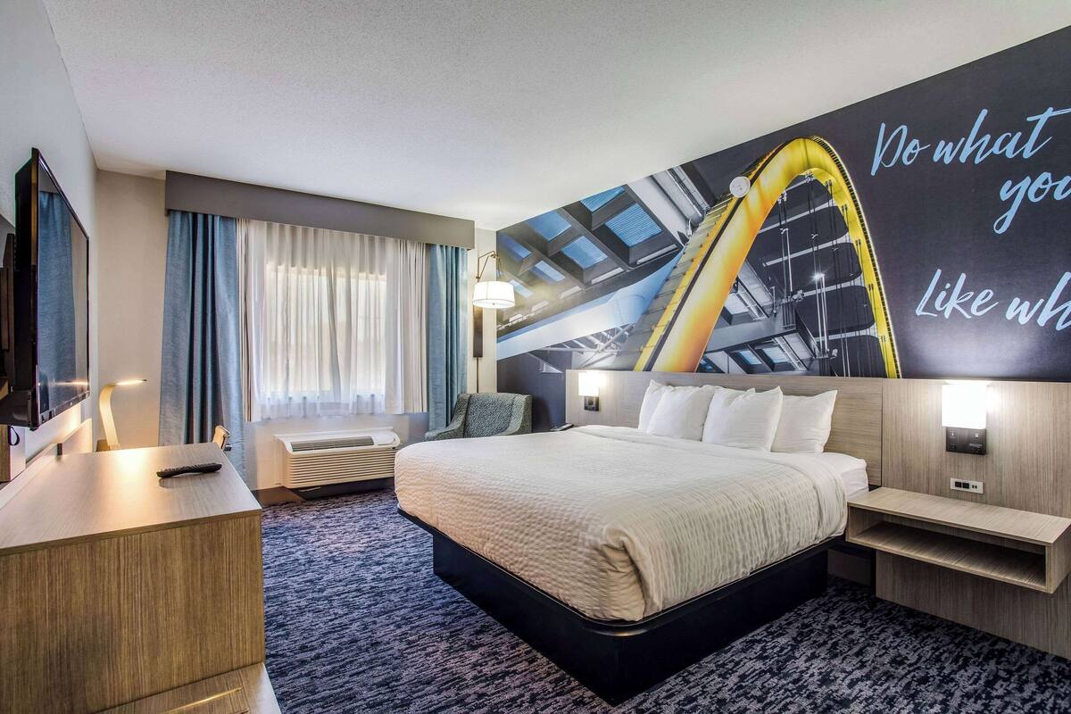 A modern hotel room with a large bed, a desk, a flat-screen TV, and a wall with motivational text.