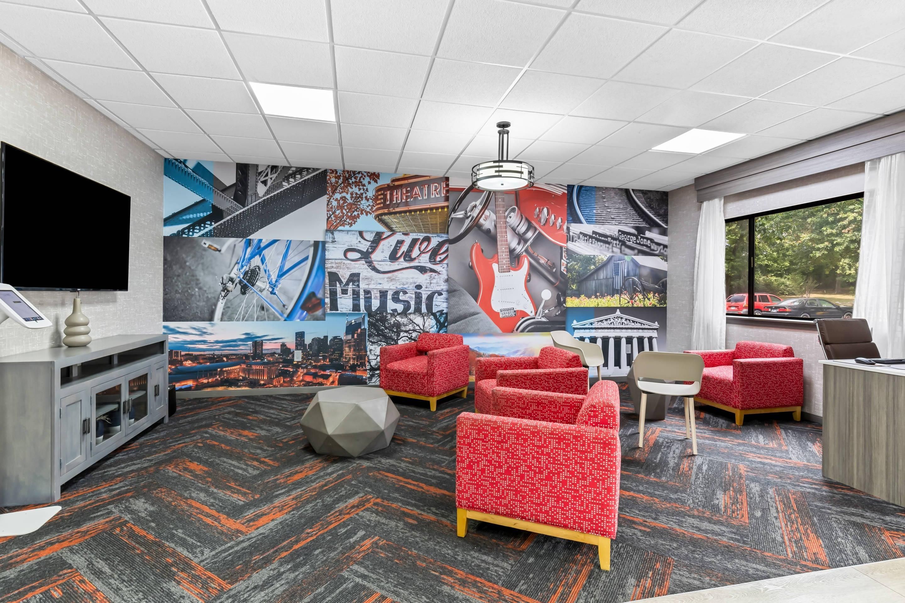 A hotel room with a cityscape mural, red patterned sofas, a desk, and a television.