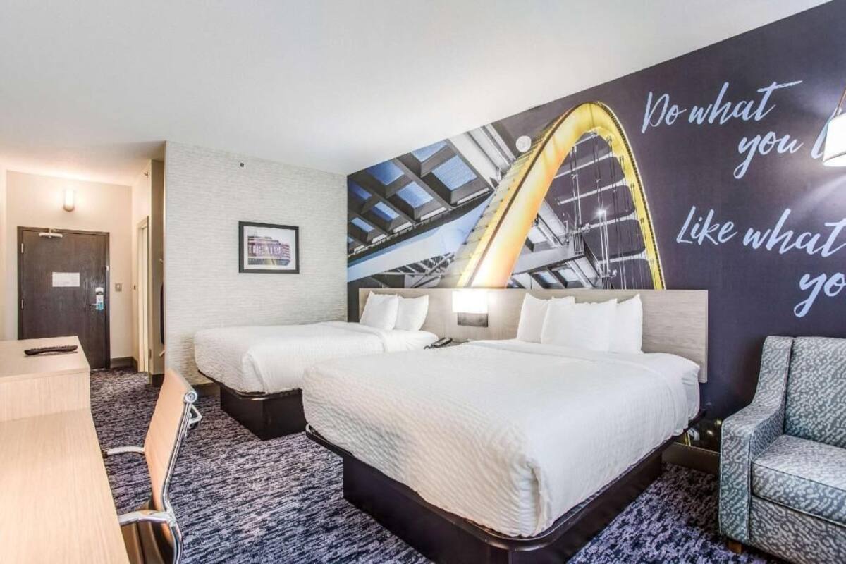A hotel room with two beds, a desk, a chair, a large wall mural, and a door.
