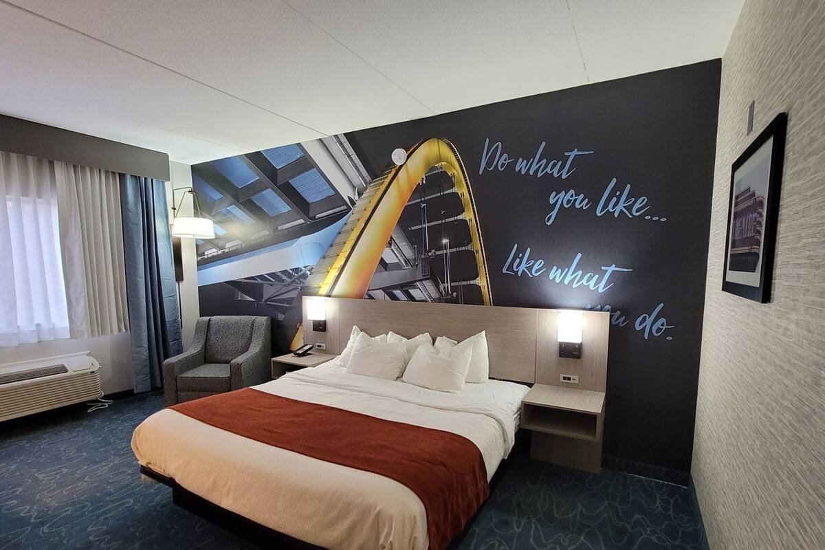 A hotel room with a bed, a desk, a chair, a wall with a motivational quote, and a window with curtains.