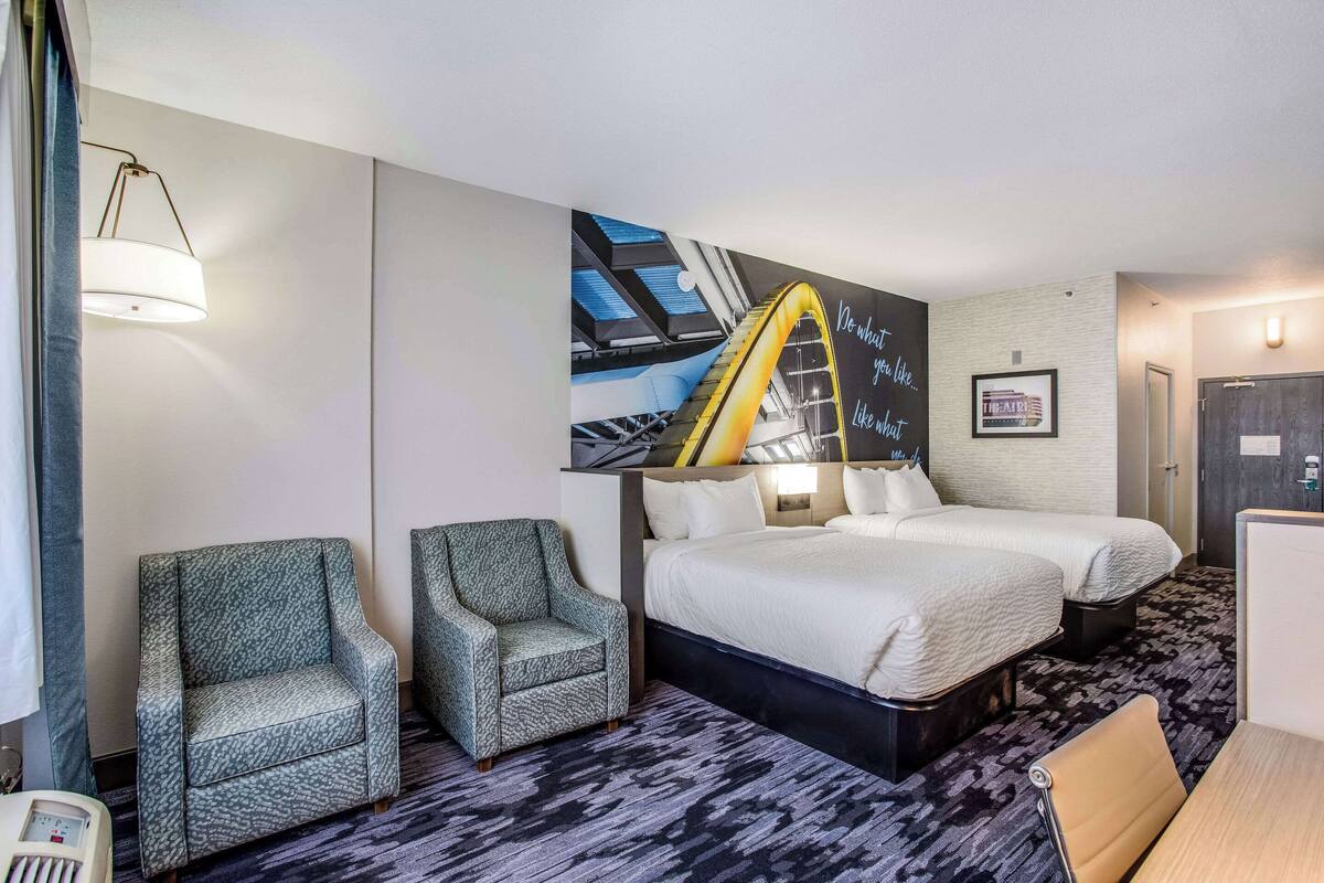 A hotel room with two beds, a large wall mural, a desk, and two armchairs.