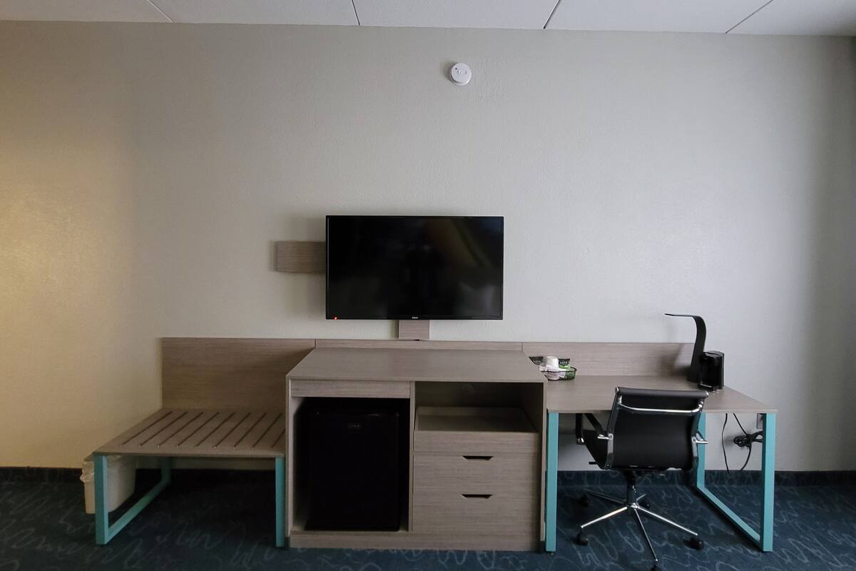 A hotel room with a desk, a chair, a television mounted on the wall, and a small trash bin.