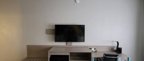A hotel room with a desk, a chair, a television mounted on the wall, and a small trash bin.