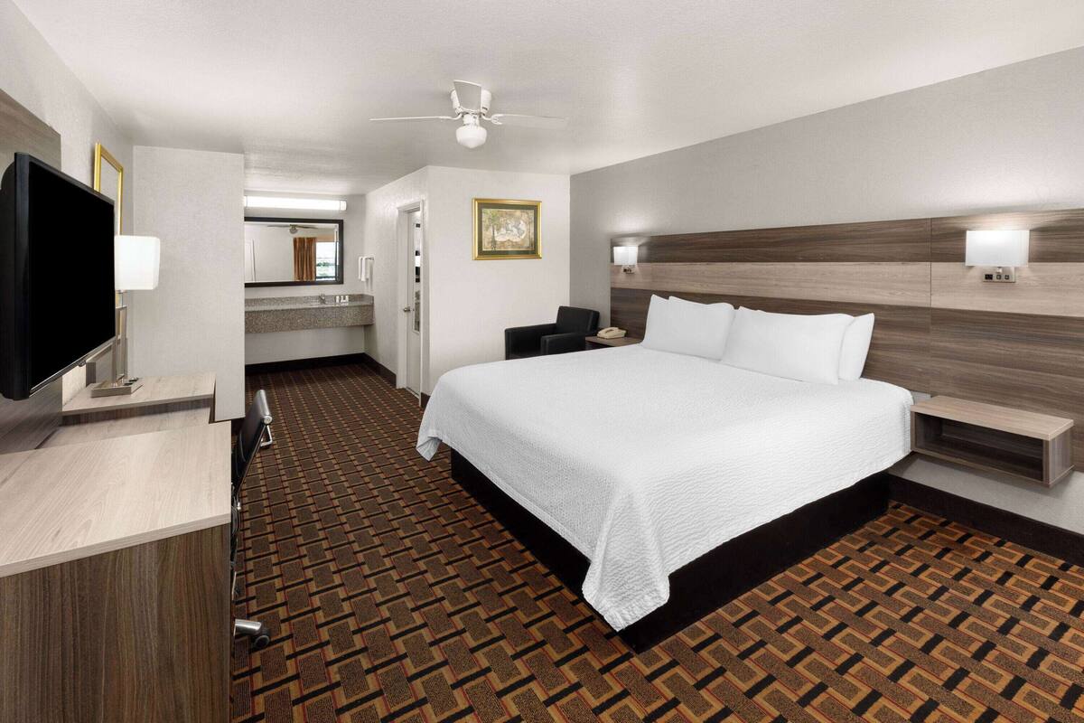 A hotel room with a bed, bedside tables, a TV, a ceiling fan, a bathroom with a mirror, and a patterned carpet.