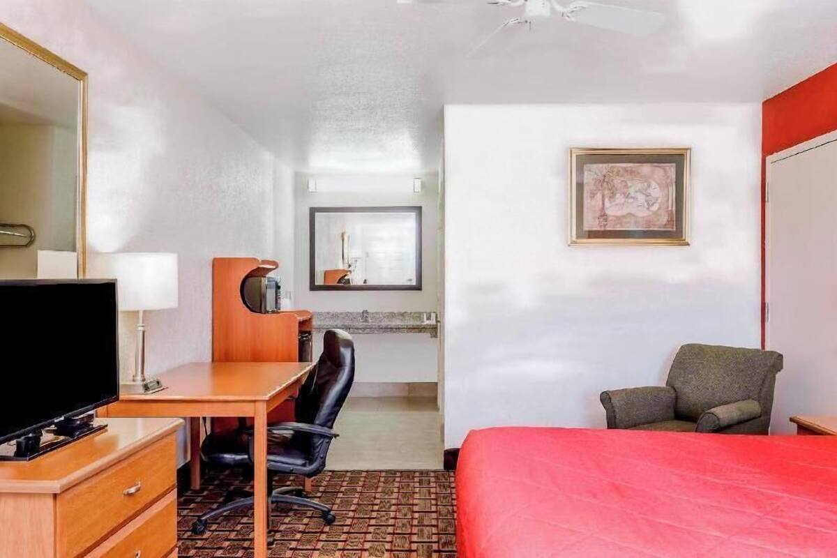 A hotel room with a bed, a desk with a chair, a television, a mirror, and a framed picture on the wall.