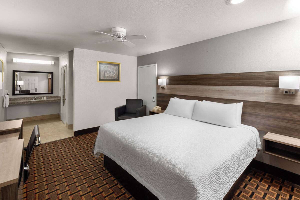 A hotel room with a large bed, a desk, a chair, a ceiling fan, a wall-mounted lamp, and a framed picture on the wall.