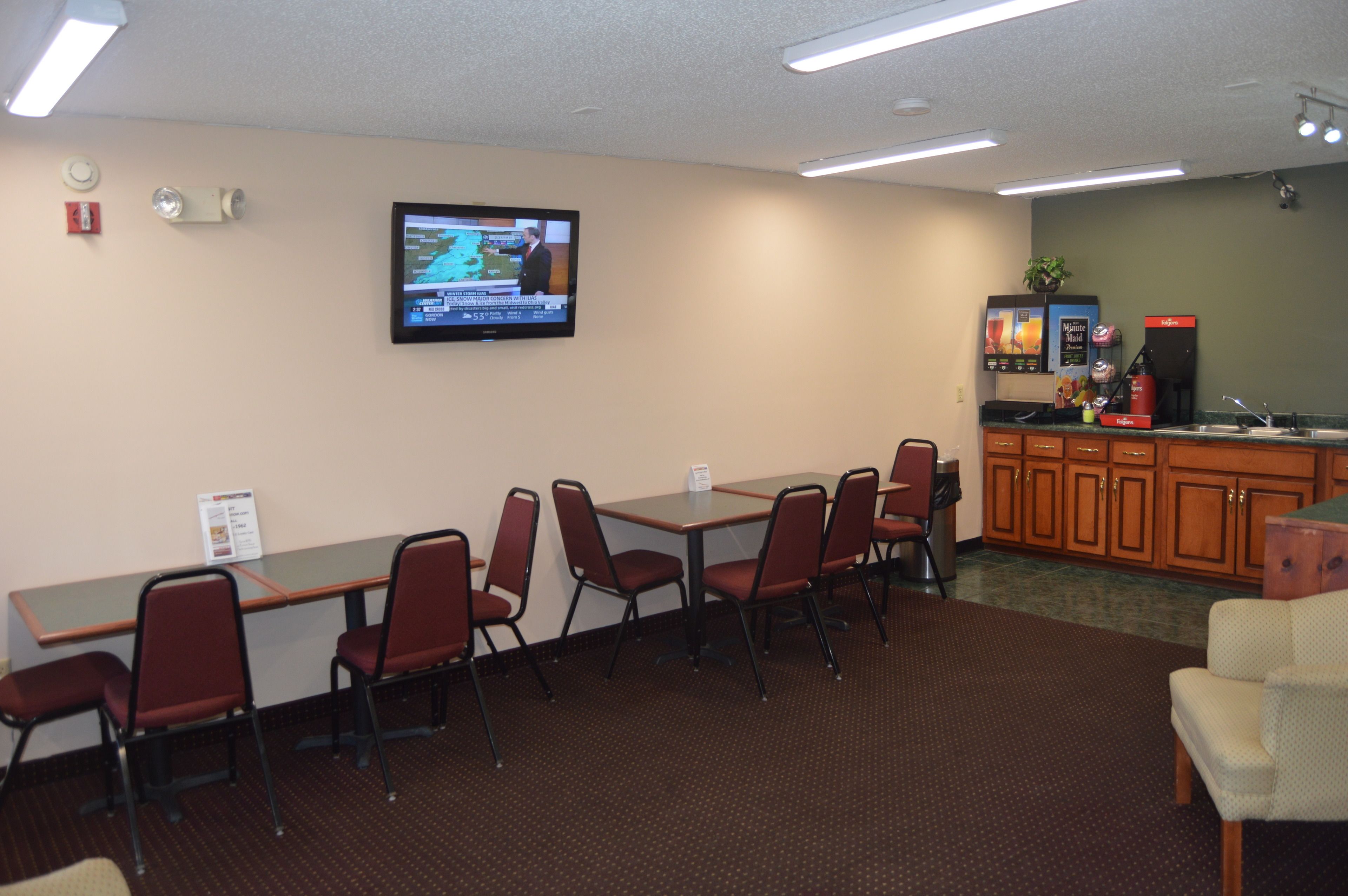 A conference room with a long table, six chairs, a television mounted on the wall, and a kitchenette area with a coffee machine and sink.