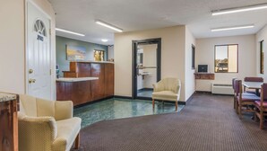Un hall d’hôtel comprenant un comptoir d’accueil, un espace d’attente avec des fauteuils, un coin repas avec une table et des chaises, et un grand miroir au mur.