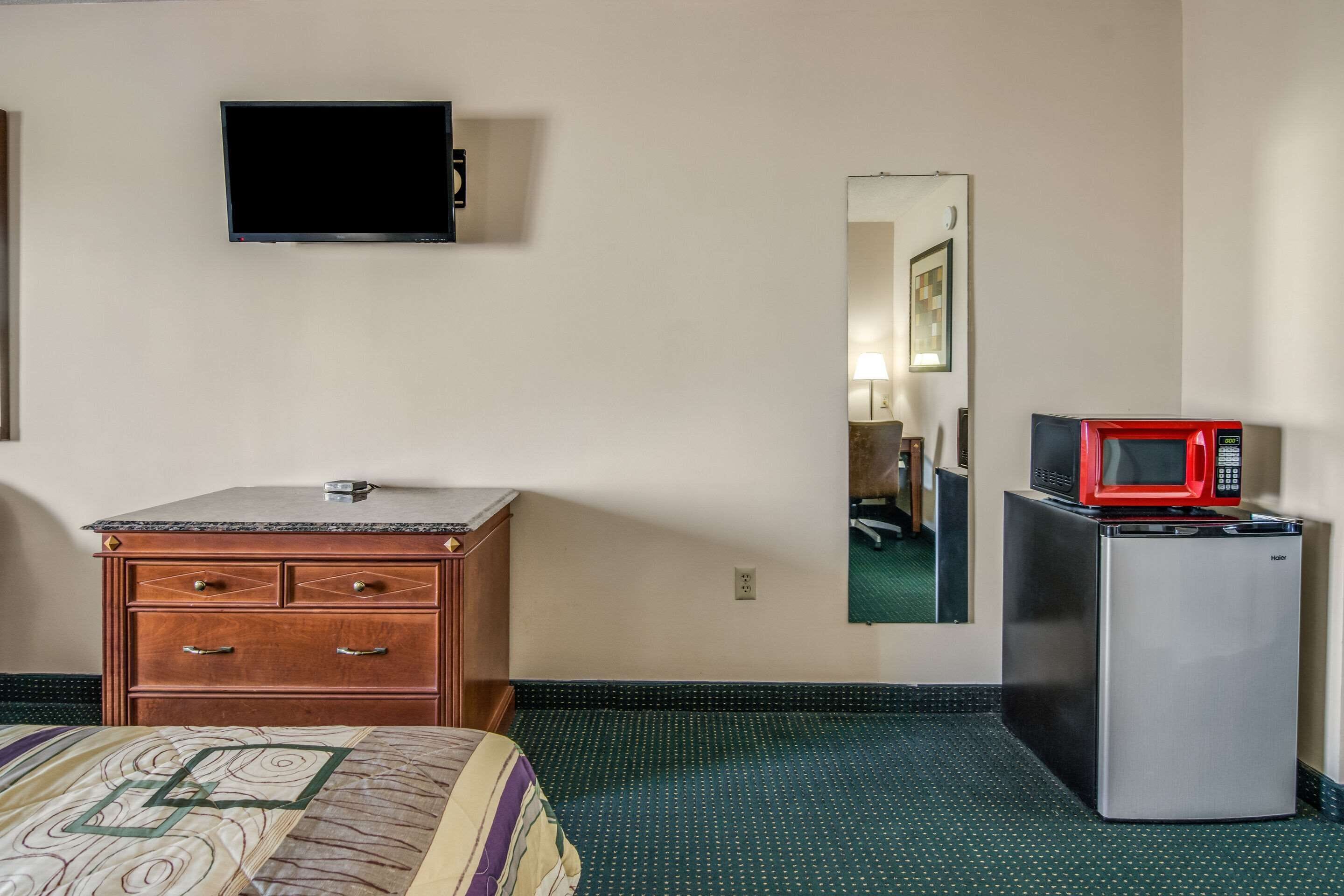 A hotel room with a bed, a wooden dresser, a television, a mirror, a refrigerator, and a microwave.