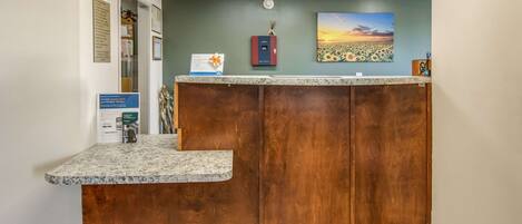 A reception desk with a wooden panel and a marble counter, a clock, and a painting of a sunset on the wall.