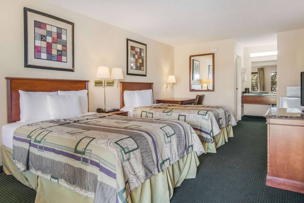 Quarto de hotel com duas camas, uma escrivaninha, uma cadeira, televisão e banheiro visível por uma porta aberta.