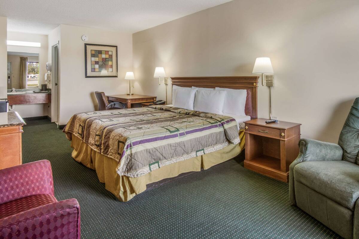 Quarto de hotel com uma cama grande, duas mesas de cabeceira com luminárias, uma escrivaninha com cadeira, um armário pequeno de madeira e uma poltrona vermelha.