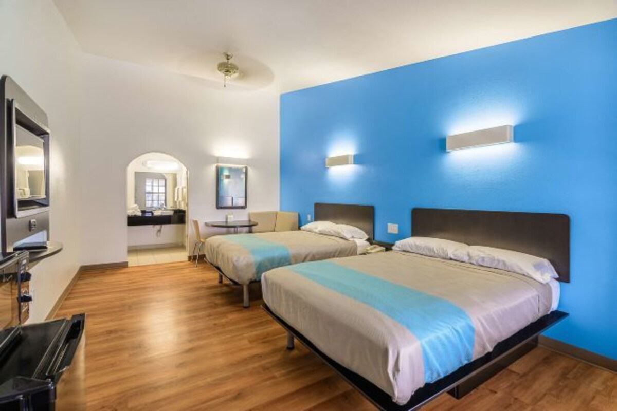 Habitación de hotel con dos camas, un televisor, un escritorio y un baño visible a través de un arco.