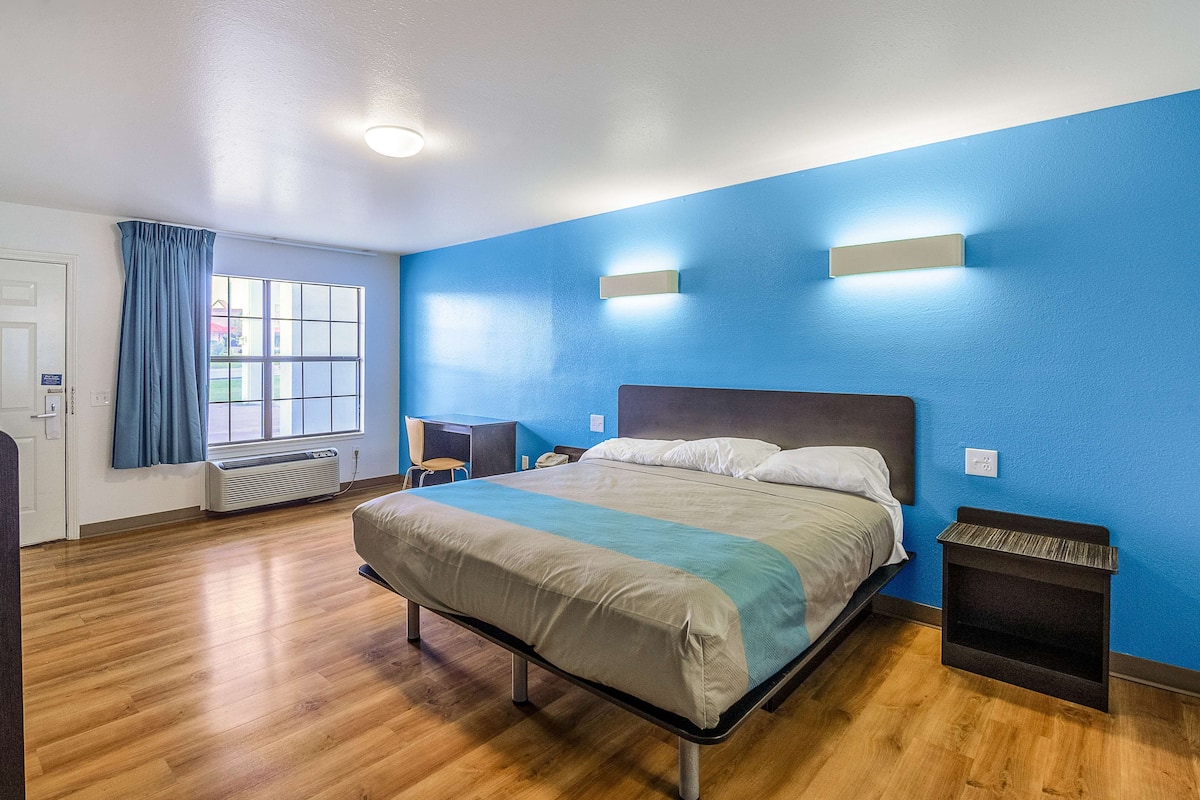 Habitación de hotel con cama, escritorio, silla, ventana con cortinas y lámpara fijada a la pared.