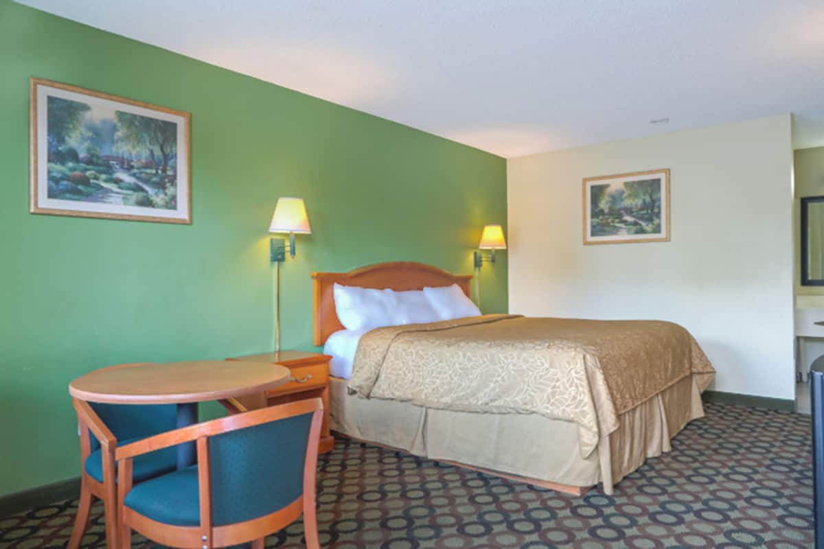 Habitación de hotel con una cama, dos lámparas de noche, una mesita redonda con una silla y dos cuadros en las paredes.