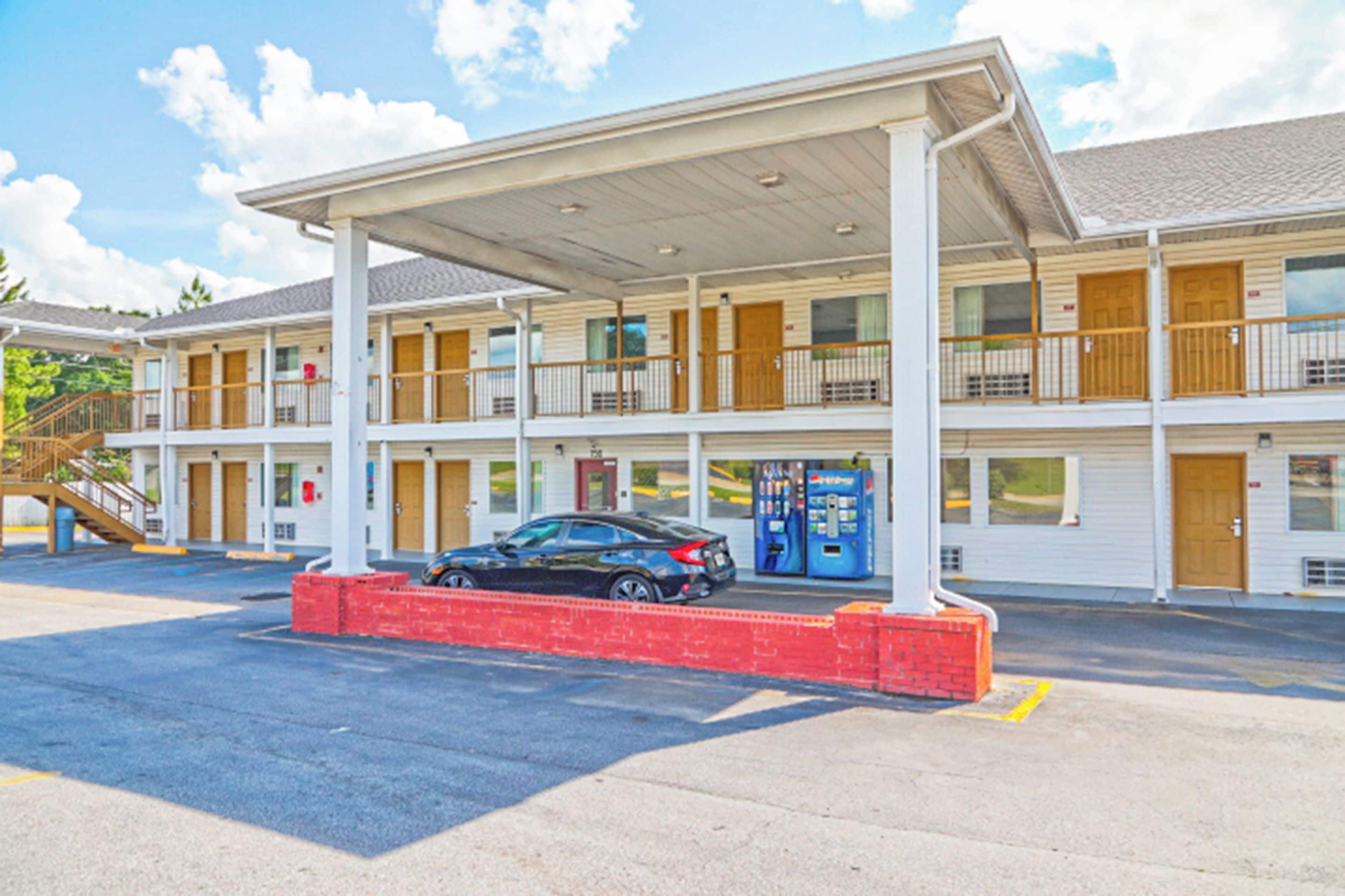Een motel met een overdekte parkeerplaats, meerdere kamers en een auto ervoor geparkeerd.