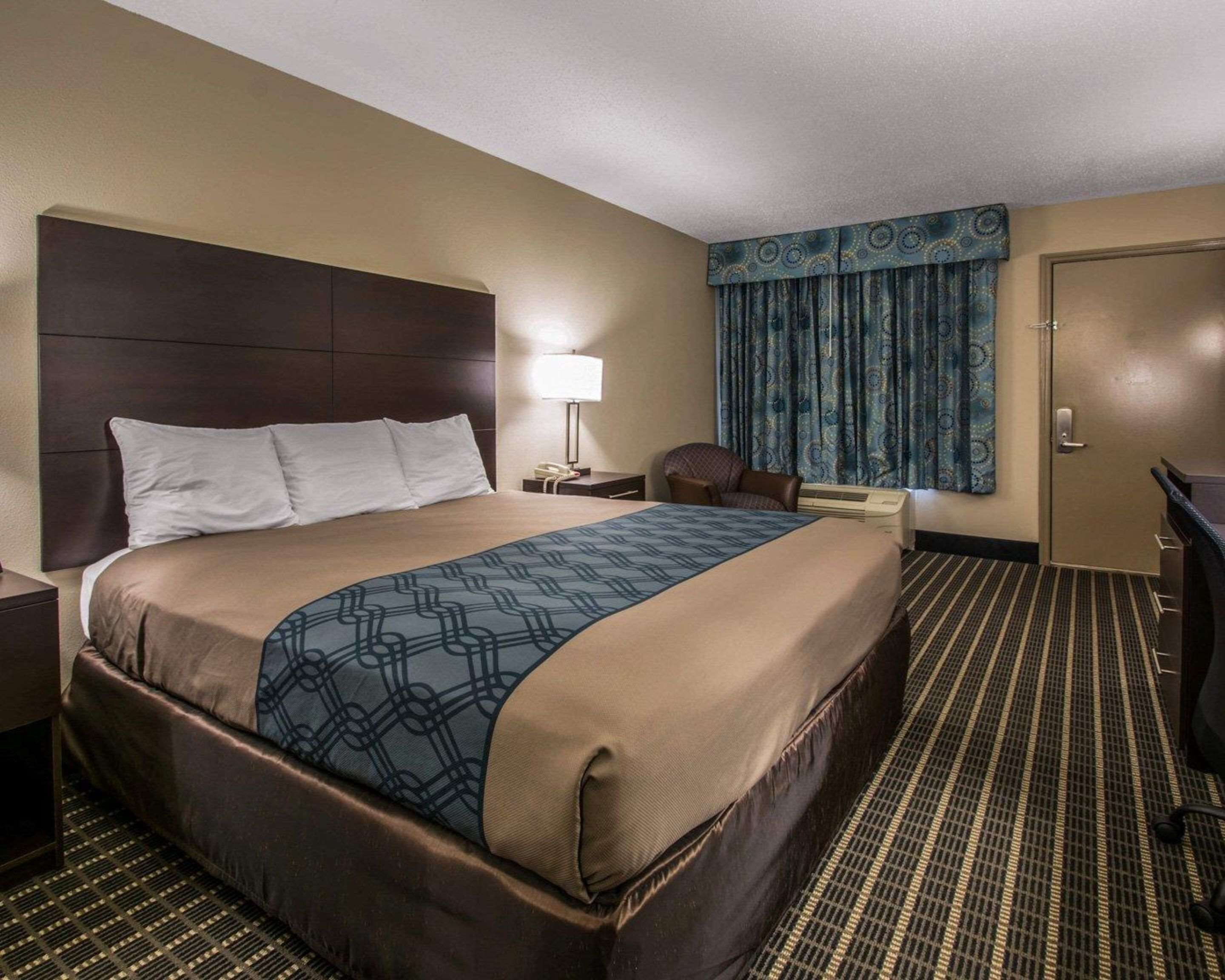 A hotel room with a large bed, a desk, a chair, a lamp, and a door.