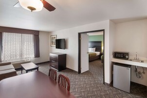 A hotel room with a dining table, chairs, a TV, a kitchenette, and a bed in the adjacent room.