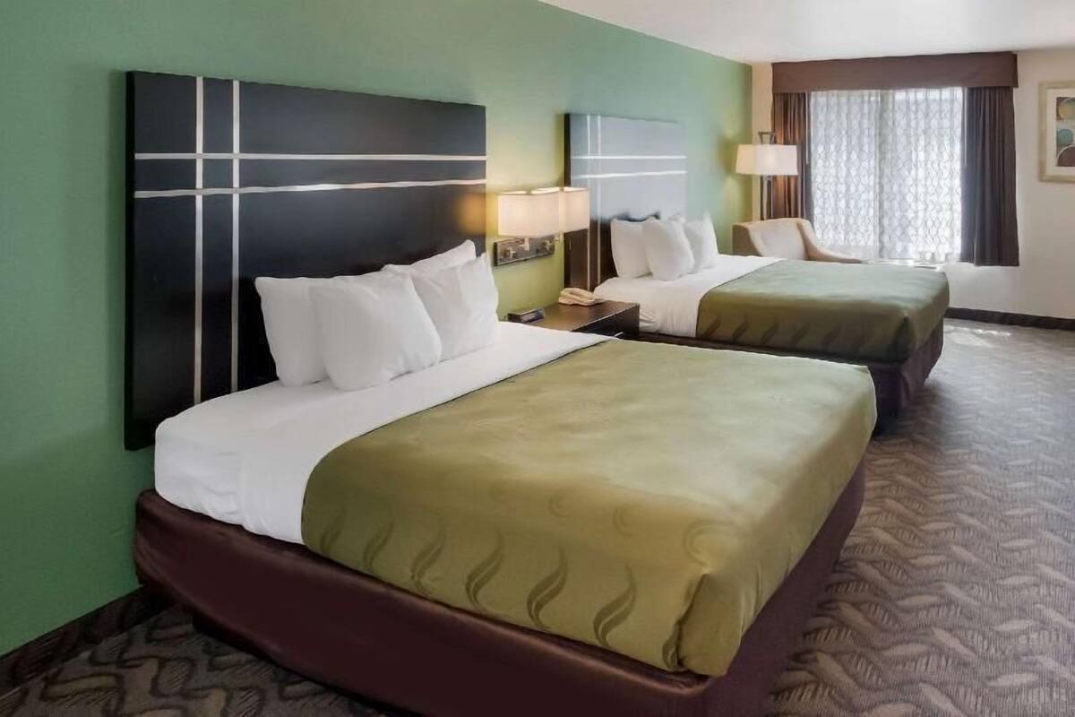 Une chambre d’hôtel avec deux lits, une tête de lit, une table de chevet, une lampe et une fenêtre avec des rideaux.