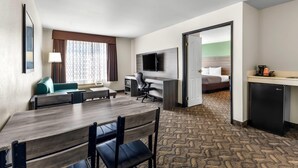 Une chambre d’hôtel comprenant un coin repas, un bureau avec une chaise, une télévision, un lit avec une tête de lit verte et une petite kitchenette.