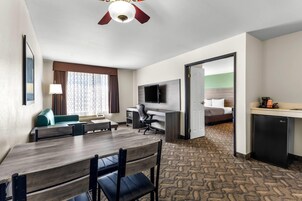 A hotel room with a dining area, a desk with a chair, a television, a bed with a green headboard, and a small kitchenette.