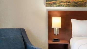 Una habitación de hotel con un sillón azul, una mesita de noche con lámpara y un cuadro de un paisaje forestal en la pared.