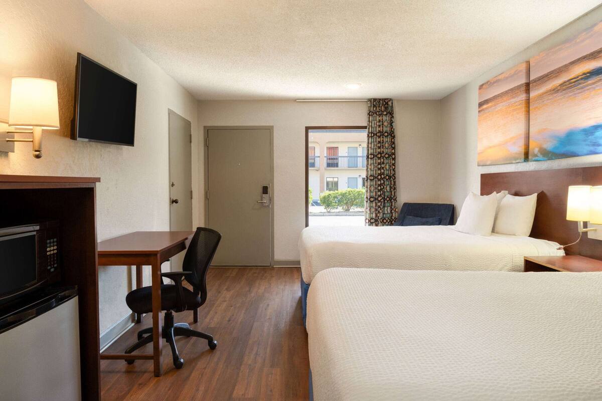 A hotel room with two beds, a desk with a chair, a TV mounted on the wall, and a painting above the bed.