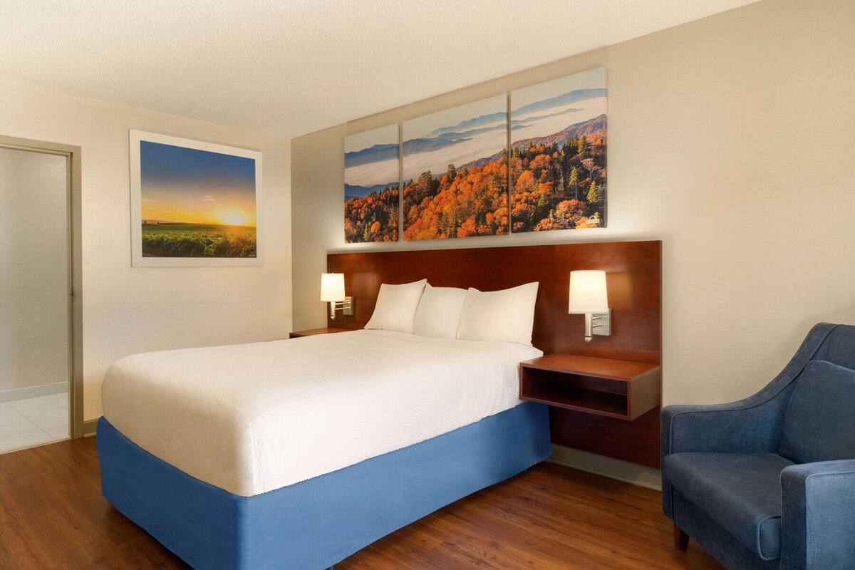 A hotel room with a large bed, a wooden headboard, two bedside lamps, a blue chair, and a painting of a sunset and autumn forest on the wall.