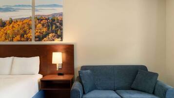 A hotel room with a bed, a blue sofa, a nightstand with a lamp, and two framed pictures of a forest landscape.