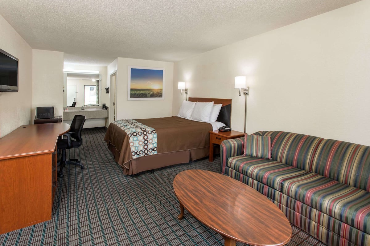 A hotel room with a bed, a sofa, a desk, a chair, a TV, and a painting on the wall.