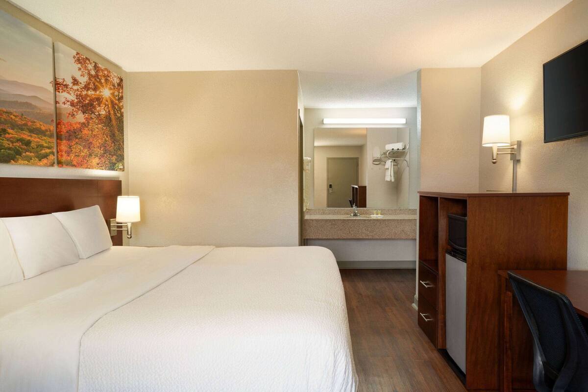 A hotel room with a large bed, a desk, a chair, a desk lamp, a mirror, and a painting on the wall.