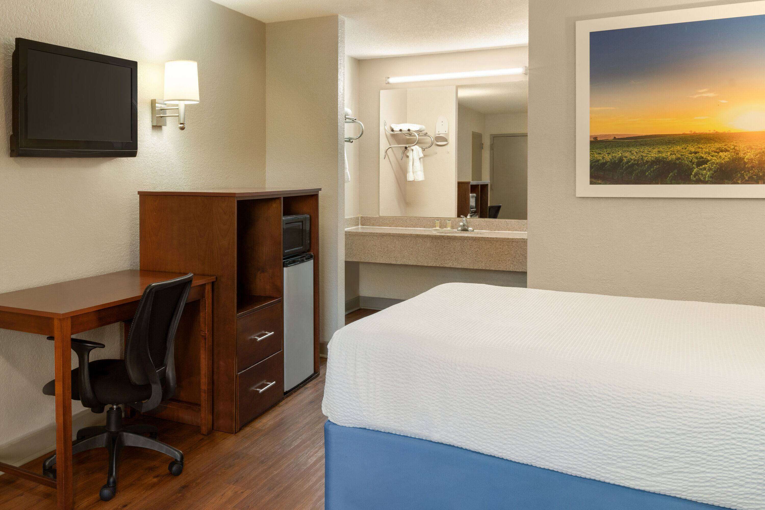 Una habitación de hotel con cama, escritorio, silla y una pequeña kitchenette.