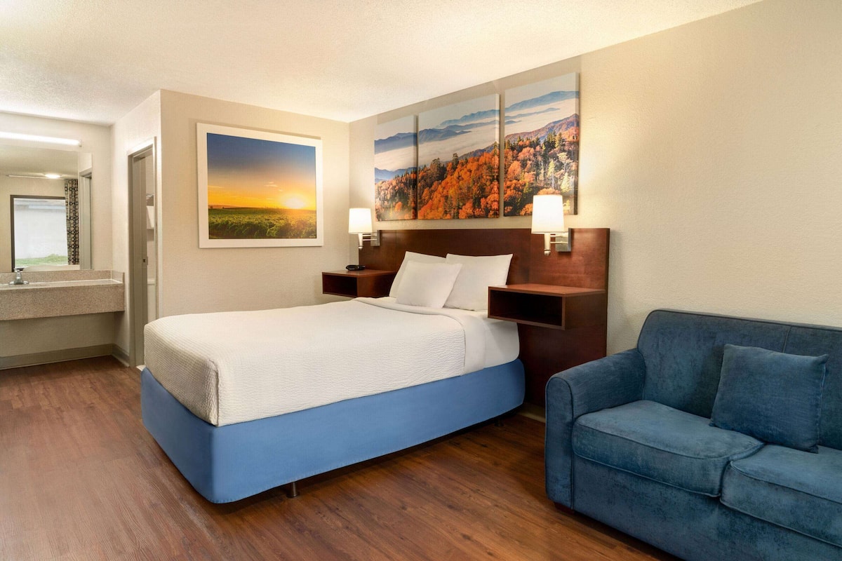 A hotel room with a bed, a blue sofa, a painting of a sunset, and a wooden headboard.
