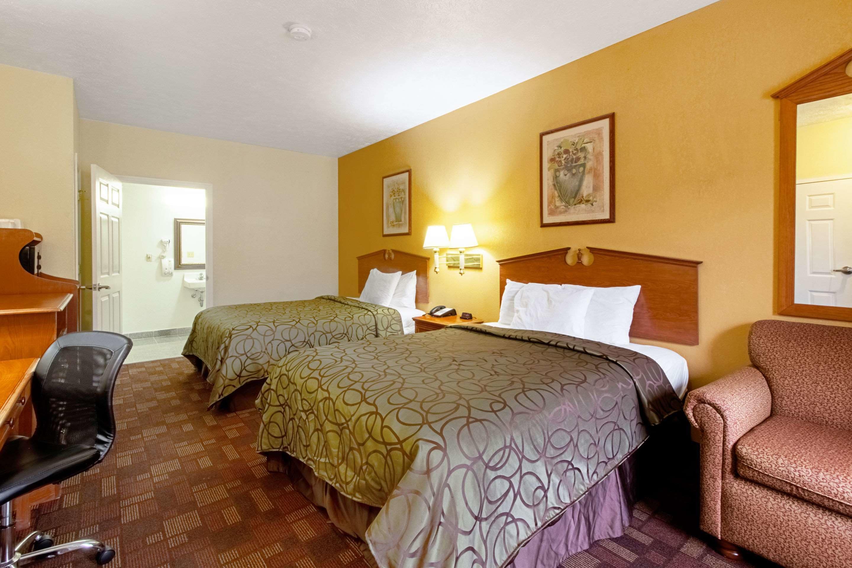 A hotel room with two beds, a desk, a chair, and a nightstand with a lamp.