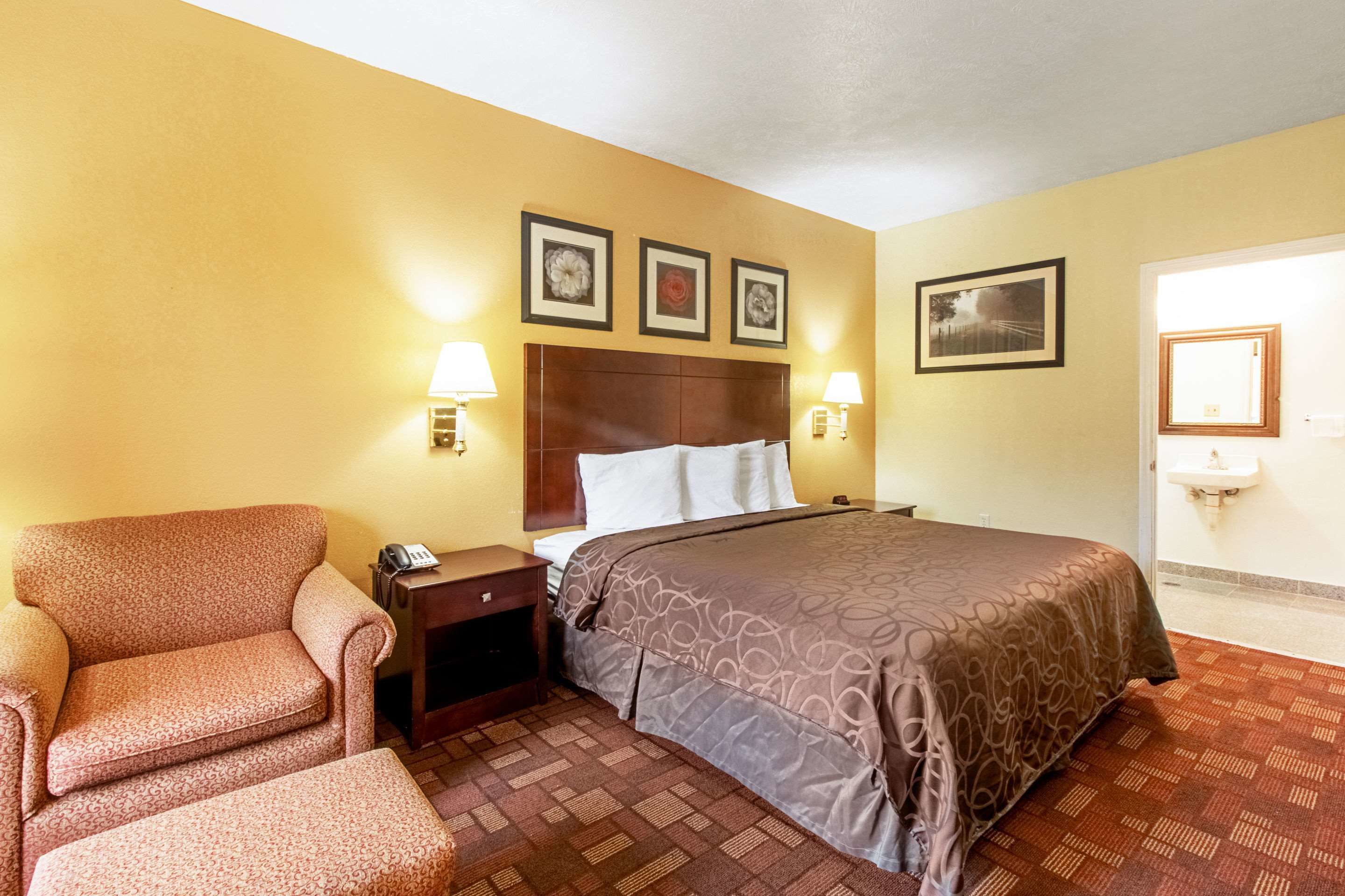 A hotel room with a bed, a chair, a nightstand, and a wall-mounted sink.