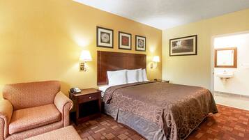 Quarto de hotel com cama, cadeira, mesa de cabeceira e pia fixa na parede.