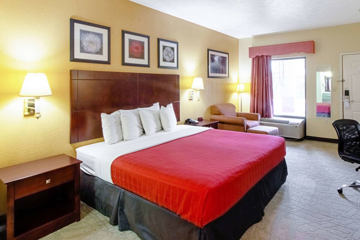 Une chambre d’hôtel avec un grand lit, deux tables de chevet, une chaise, un bureau et une fenêtre avec des rideaux.