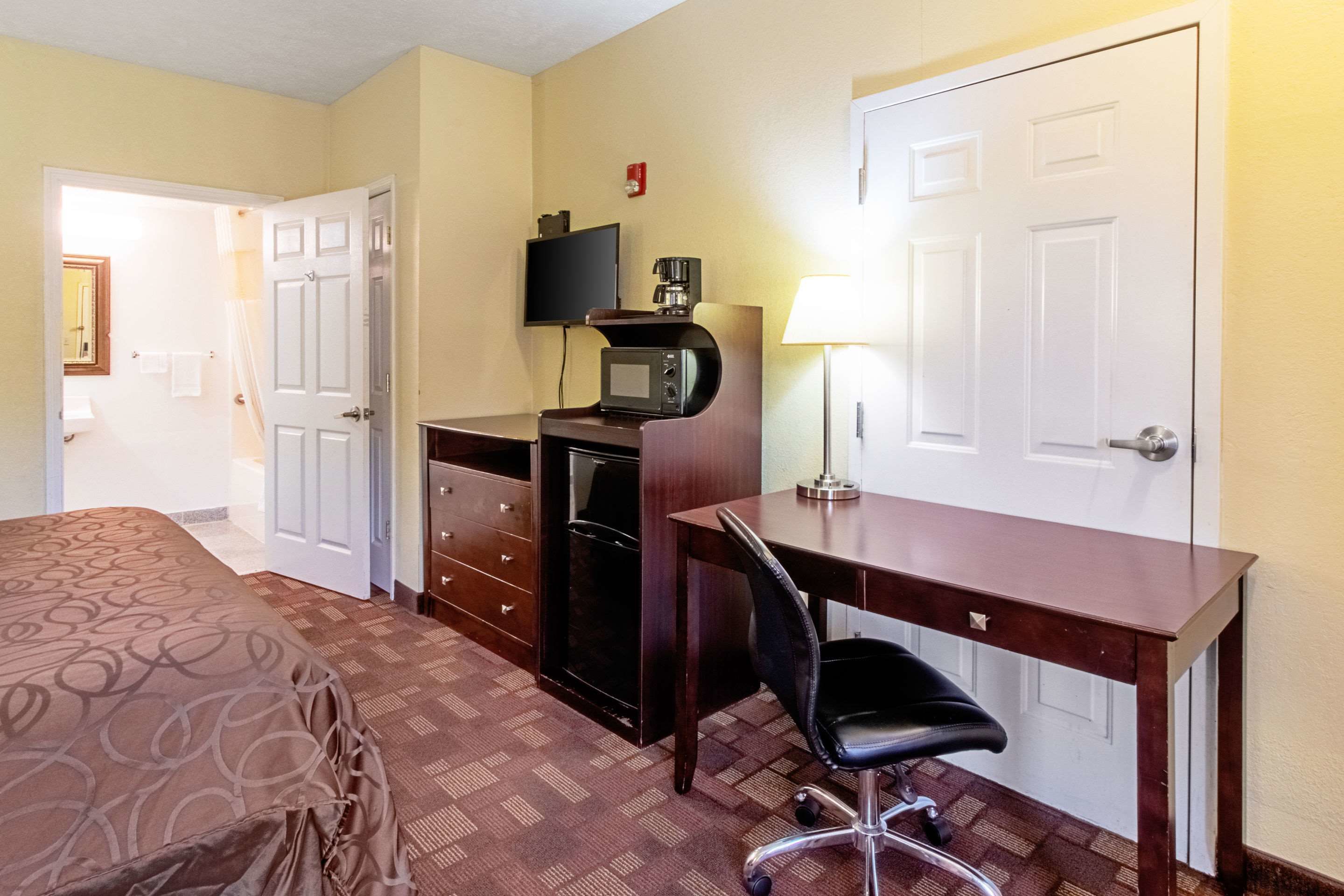 A hotel room with a bed, a desk with a chair, a television, a coffee maker, and a lamp.