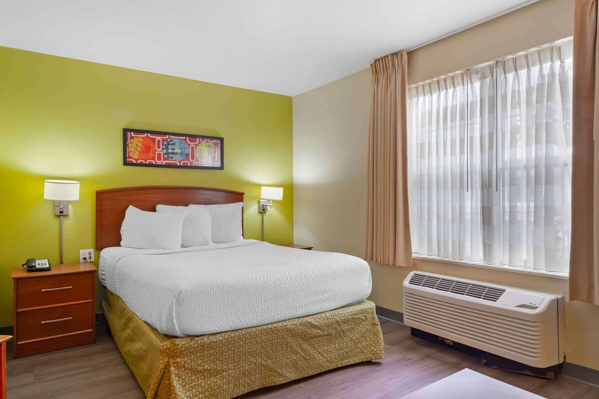 A hotel room with a bed, bedside lamps, a nightstand, a heater, and a window with curtains.