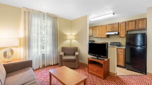 A compact hotel room with a sofa, a TV, a small table, a lamp, a chair, a kitchen area with a fridge, microwave, and wooden cabinets. - Extended Stay America Suites Atlanta Norcross Peachtree Corn (Norcross)