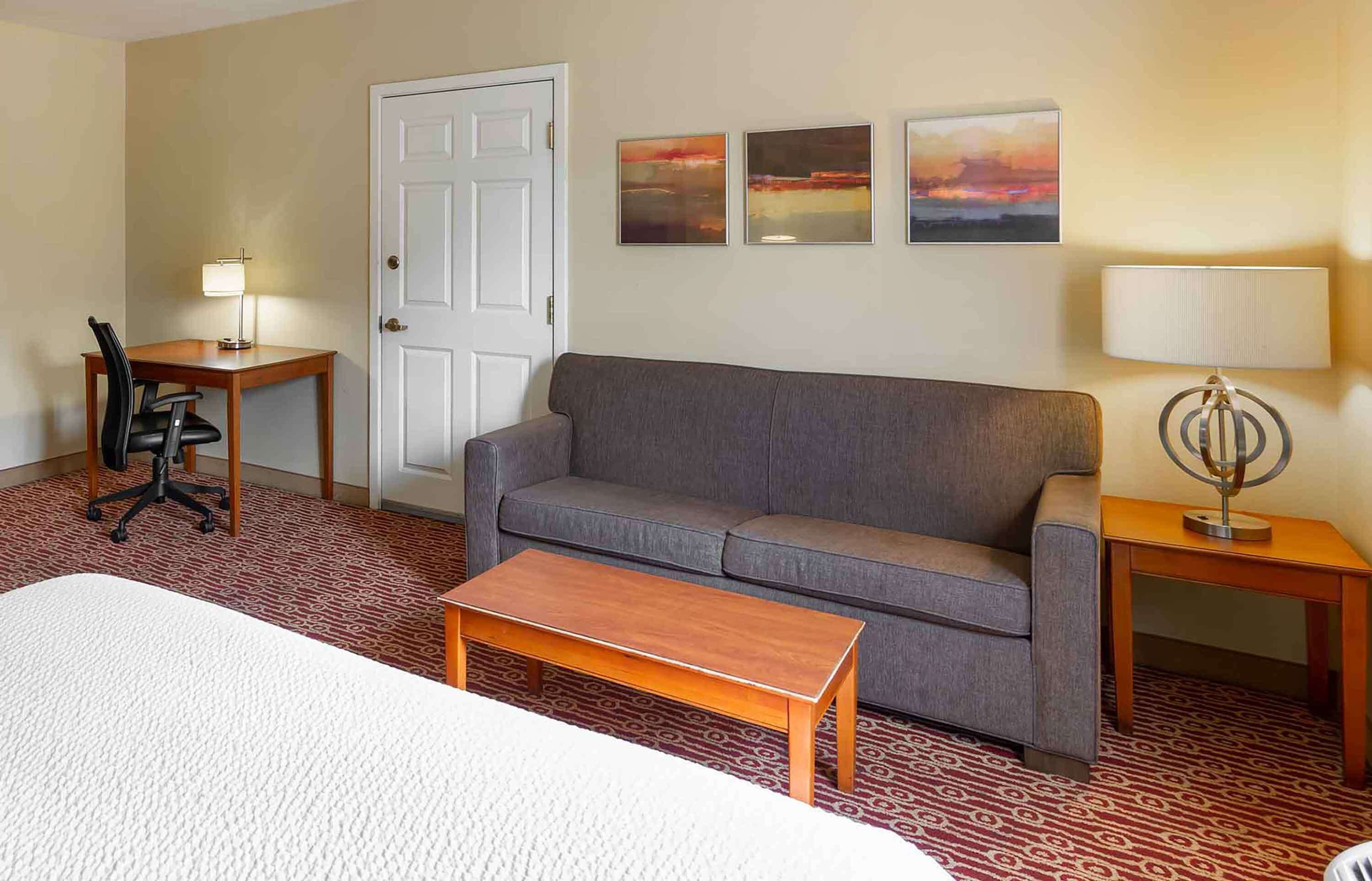 A hotel room with a gray sofa, a wooden coffee table, a desk with a chair, a lamp, and three framed pictures on the wall.