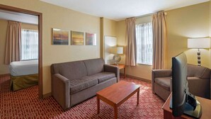A hotel room with a sofa, a coffee table, a TV, and a bed. There are two windows with curtains, a wall-mounted lamp, and a painting above the sofa. - Extended Stay America Suites Atlanta Norcross Peachtree Corn (Norcross)