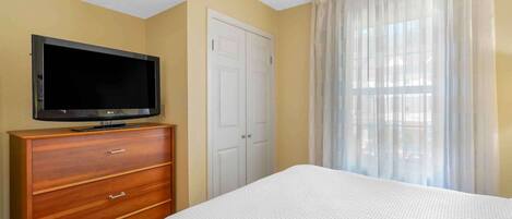 A hotel room with a bed, a TV on a wooden dresser, a white door, and a window with sheer curtains.