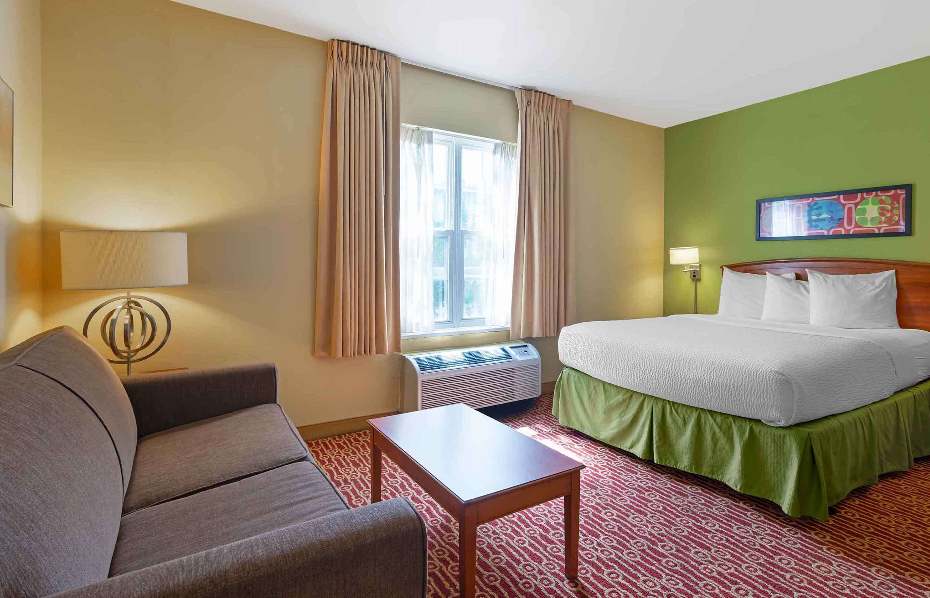 Une chambre d’hôtel comprenant un lit, un canapé, une table, une fenêtre avec des rideaux et une lampe fixée au mur.