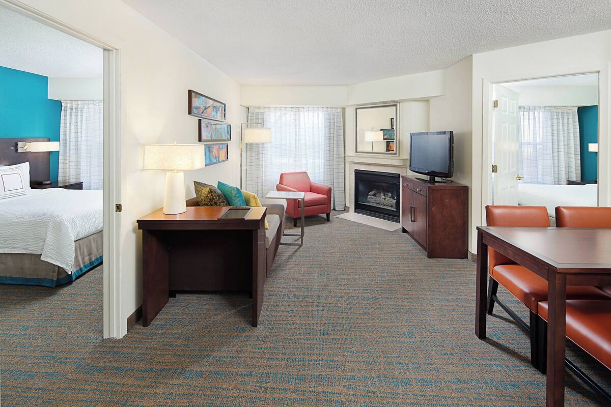 A hotel room with a bed, a desk with a lamp, a red chair, a television, and a fireplace.