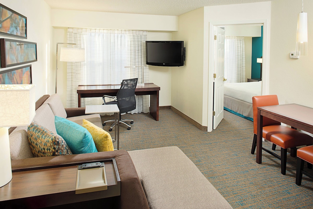 A hotel room with a bed, a desk with a chair, a TV mounted on the wall, and a dining table with chairs.