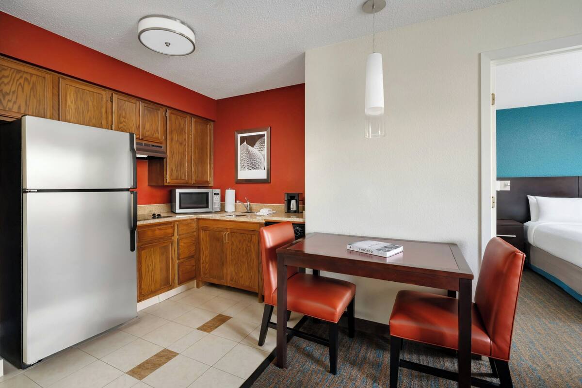 A compact hotel room with a kitchenette, a dining area with a table and chairs, a bed, and a single hanging light fixture.
