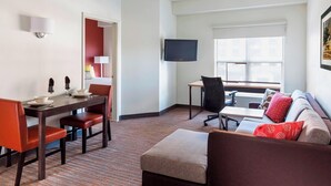 Habitació d'hotel amb zona de menjador, un televisor fixat a la paret, un sofà amb coixinets vermells i una finestra amb persianes.