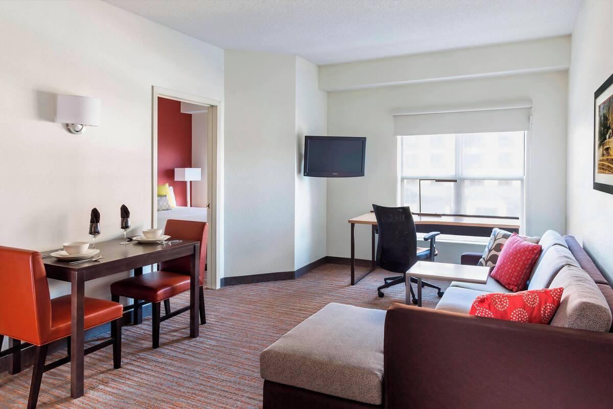 Ein Hotelzimmer mit Essbereich, einem an der Wand befestigten Fernseher, einer Couch mit roten Kissen und einem Fenster mit Jalousien.
